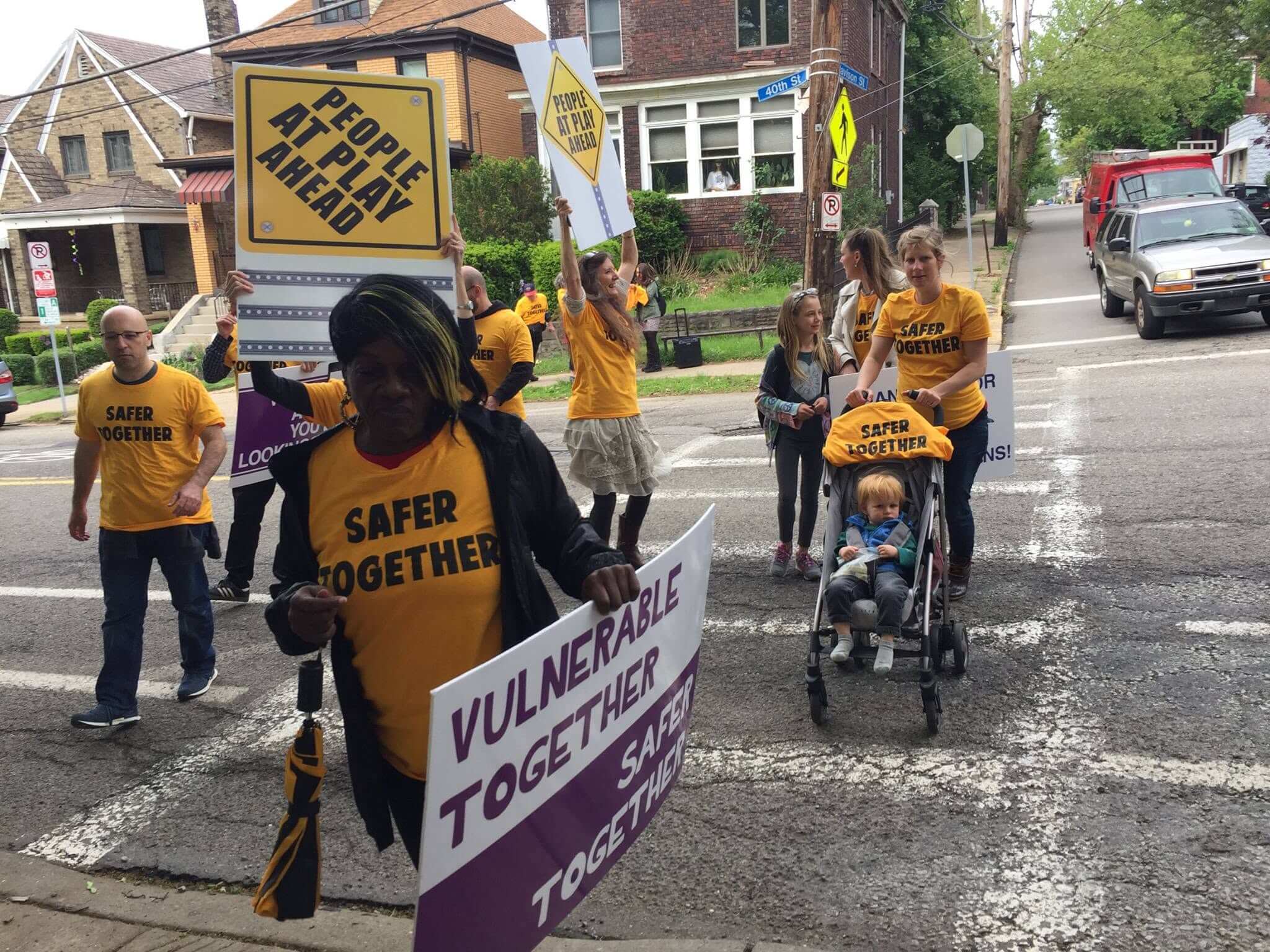 A group of people with matching shirts and signs stand in a cross walk letting others know that people are at play head.