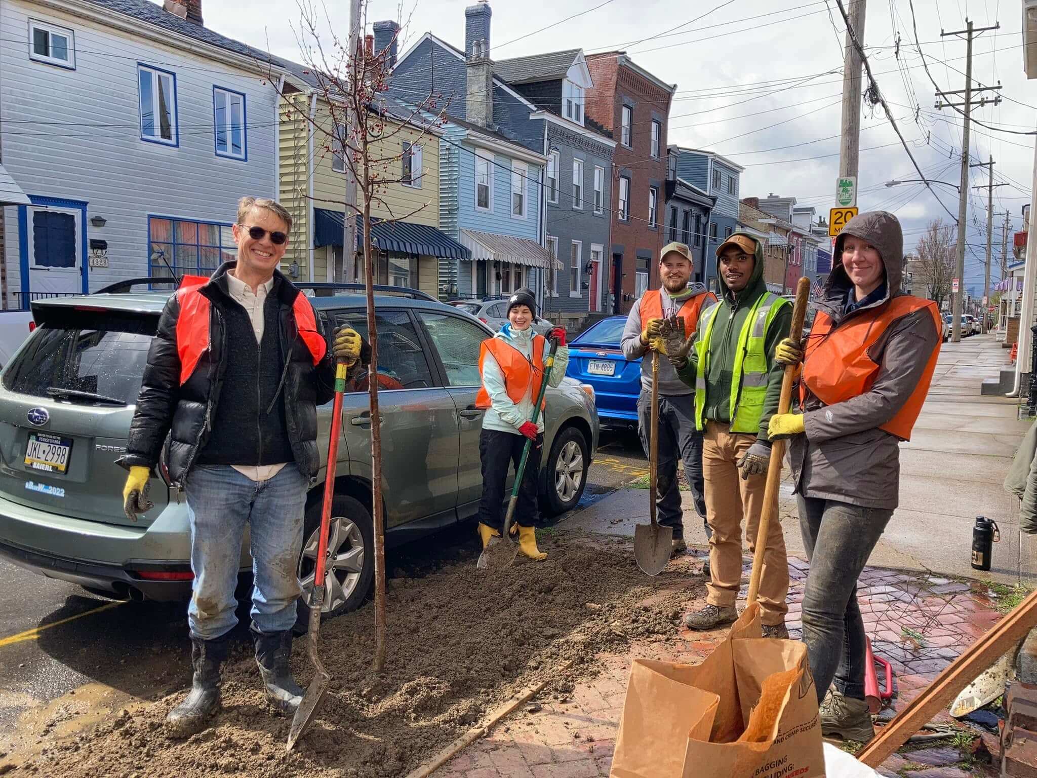 Five people pose for a photo while planting a tree in a sidewalk
